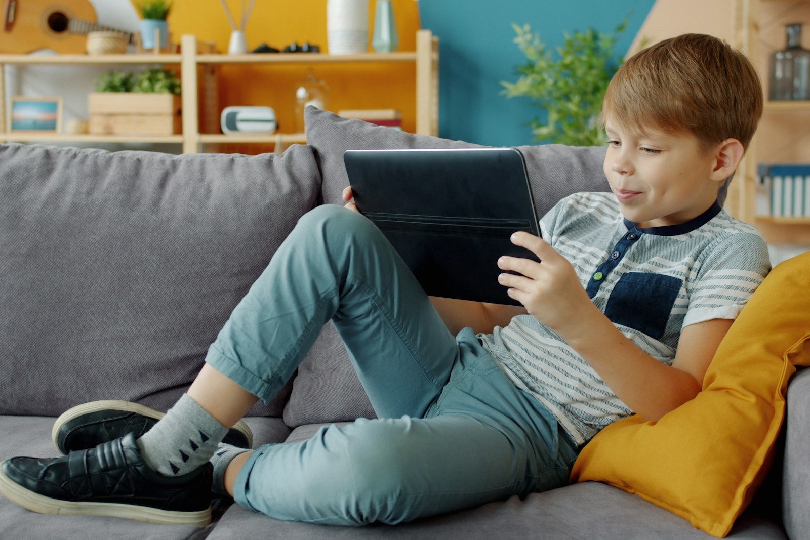 A young boy relaxing on a grey sofa with a yellow cushion, smiling at a tablet held on his knees