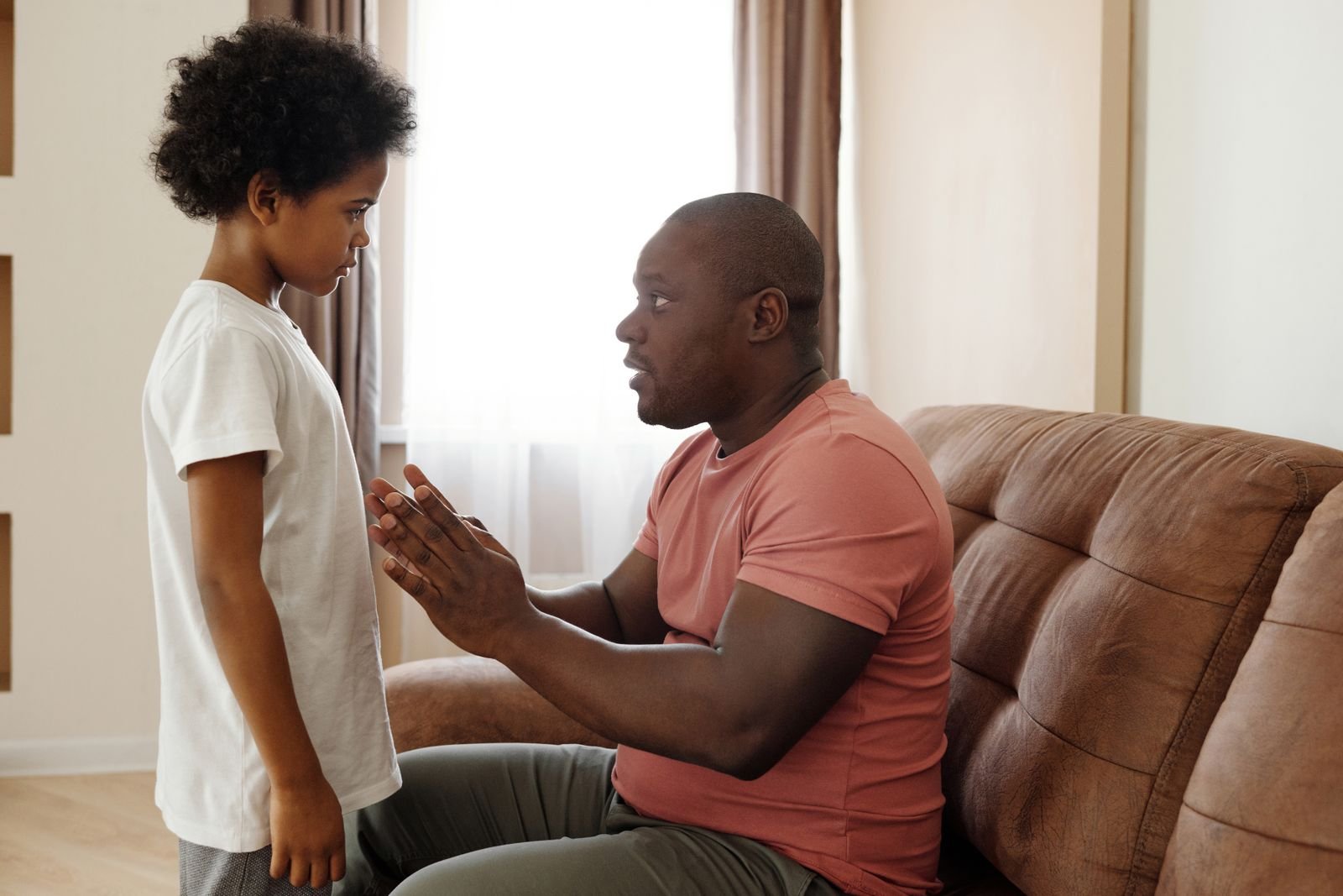 A father sitting on a sofa talking calmly with a young boy who stands beside him
