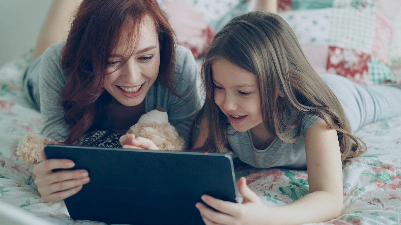A smiling mother and daughter lying on a bed, looking at a tablet together