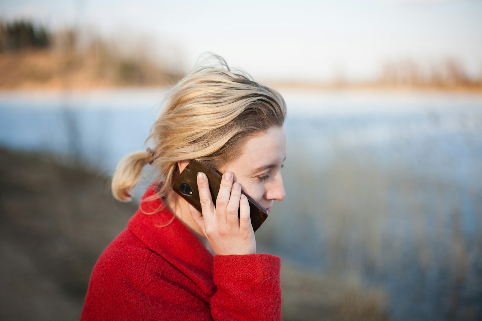 A woman in a red coat listening closely to a phone call outdoors, eyes lowered