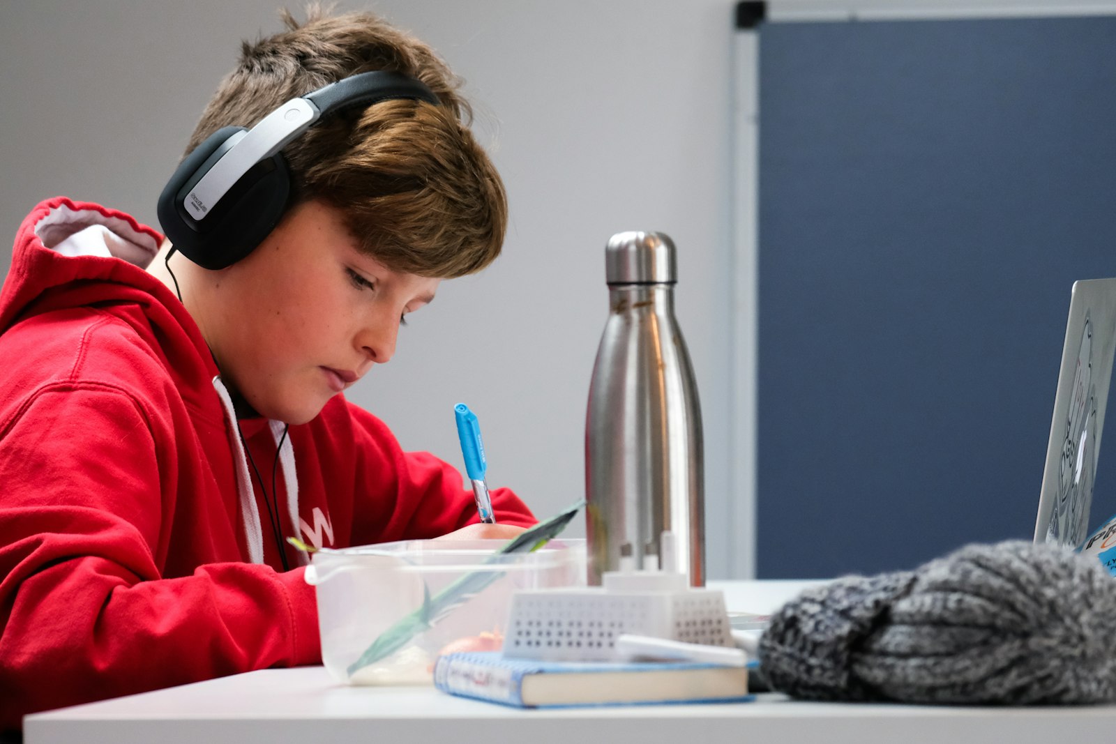 Boy wearing headphones focused on writing at a desk, laptop open beside him