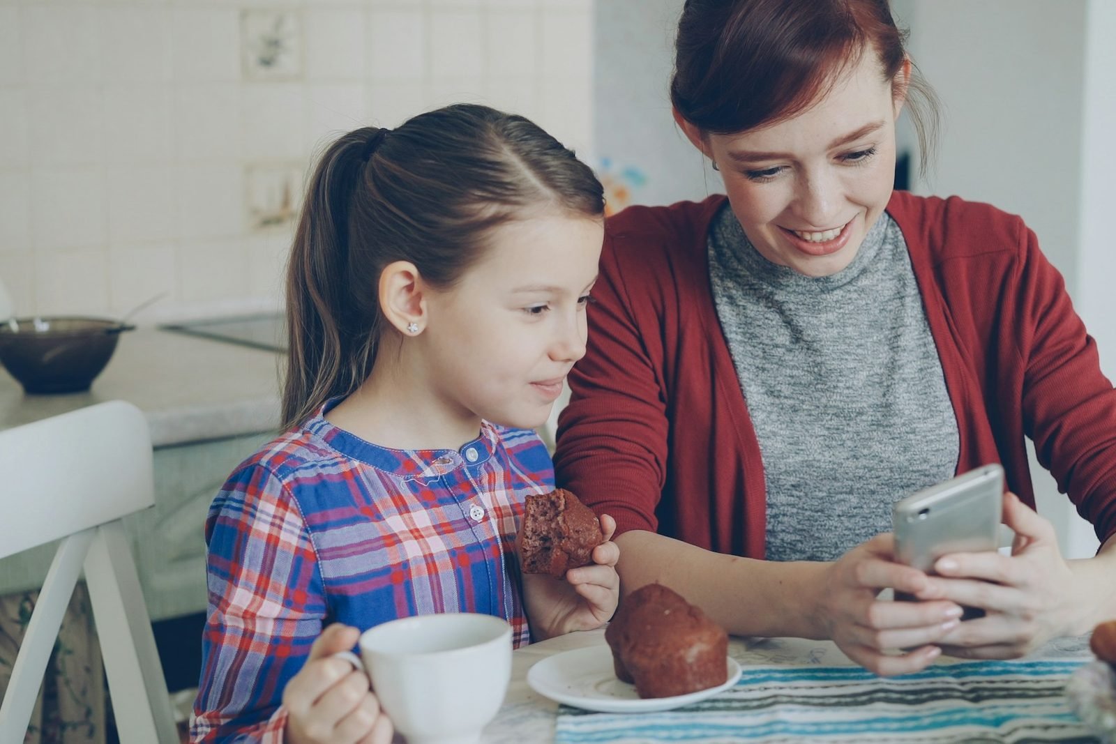 A mother and her young daughter at a kitchen table looking at a smartphone together