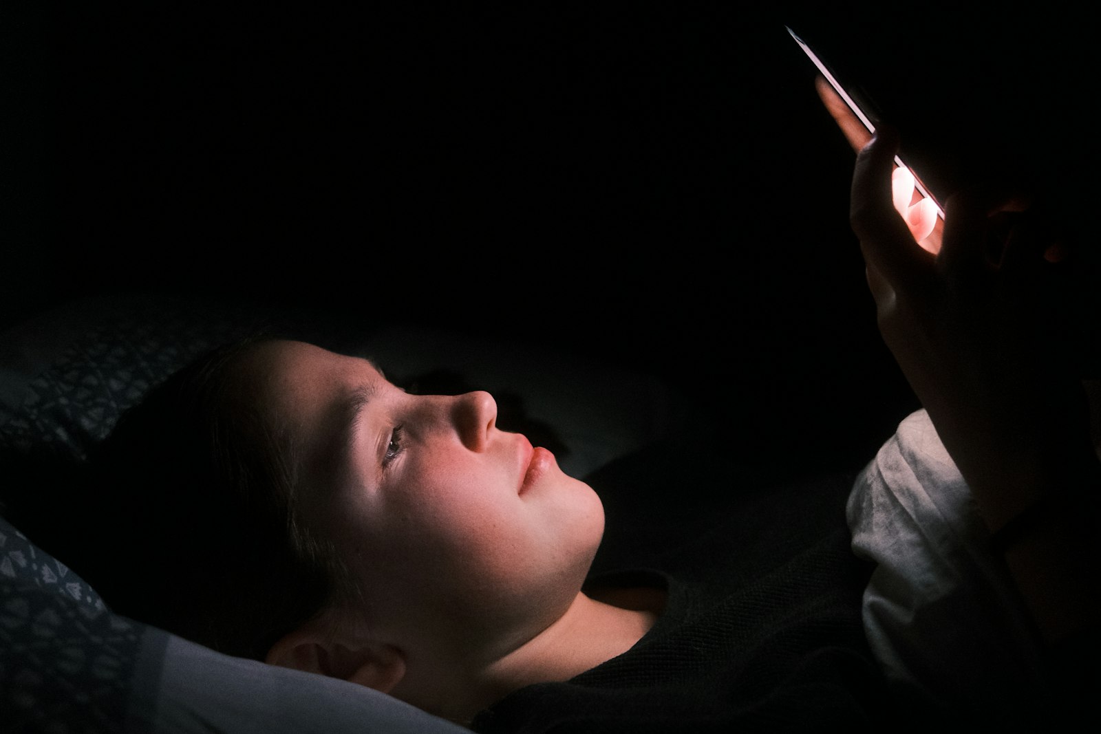 A young person lying in bed in a dark room, face lit by the glow of a phone screen